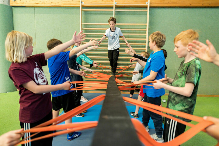 Ein Schüler geht über eine Slack-Line. Diese wird von seinen Mitschülern gehalten. Die Schüler absolvieren die Aufgabe im Rahmen eines Teambuildingmoduls im Ahorn-Sportpark.