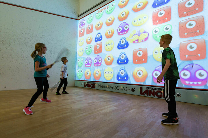 Schüler stehen mit Squash-Schlägern auf dem interaktiven Squash-Court und spielen mit Bällen an die interaktive Wand. Dort versuchen die Schüler verschiedene Monster-Icons zu treffen, um Punkte zu erzielen.