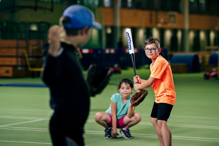 Ein Schüler holt für den Wurf des Baseballs aus. Ein anderer Schüler steht ihm mit einem Baseballschläger gegenüber und fokussiert den Ball. Hinter dem zweiten Schüler hockt eine Schülerin, um den Ball zu fangen, wenn dieser nicht vom Schüler mit dem Baseballschläger getroffen wurde.