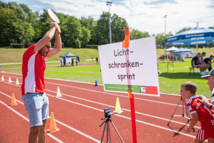 Während des Radio Hochstift-Ferienfinales hält ein Trainer vom LC Paderborn die Starklappe für den Sprintstart in die Luft. Ein Junge ist bereit loszulaufen. Um ihn herum sind Lichtschranken zur Zeitmessung des Laufes aufgebaut, um seine Schnelligkeit zu messen, und eine Rheie von Hütchen kennzeichnen die Strecke. Im Vordergrund ist eine Stange mit einem Schild aufgestellt, auf diesem steht: Lichtschrankensprint!