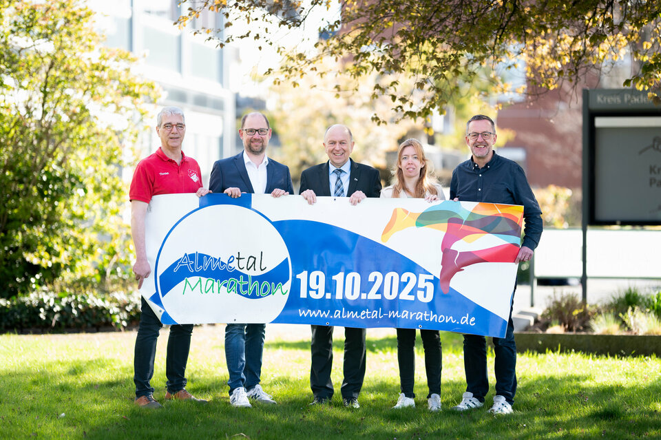 Thorsten Brinkmann (SV Rot-Weiß Alfen), Ralf Pahlsmeier (Ahorn-Sportpark), Landrat Christoph Rüther, Maren Richts (Ahorn-Sportpark) und Holger Hayn (SV Rot-Weiß Alfen) stehen mit dem Ankündigungsbanner des 8. Almetal-Marathons vor dem Kreishausgebäude. Auf dem Banner steht der 19.10.2025.