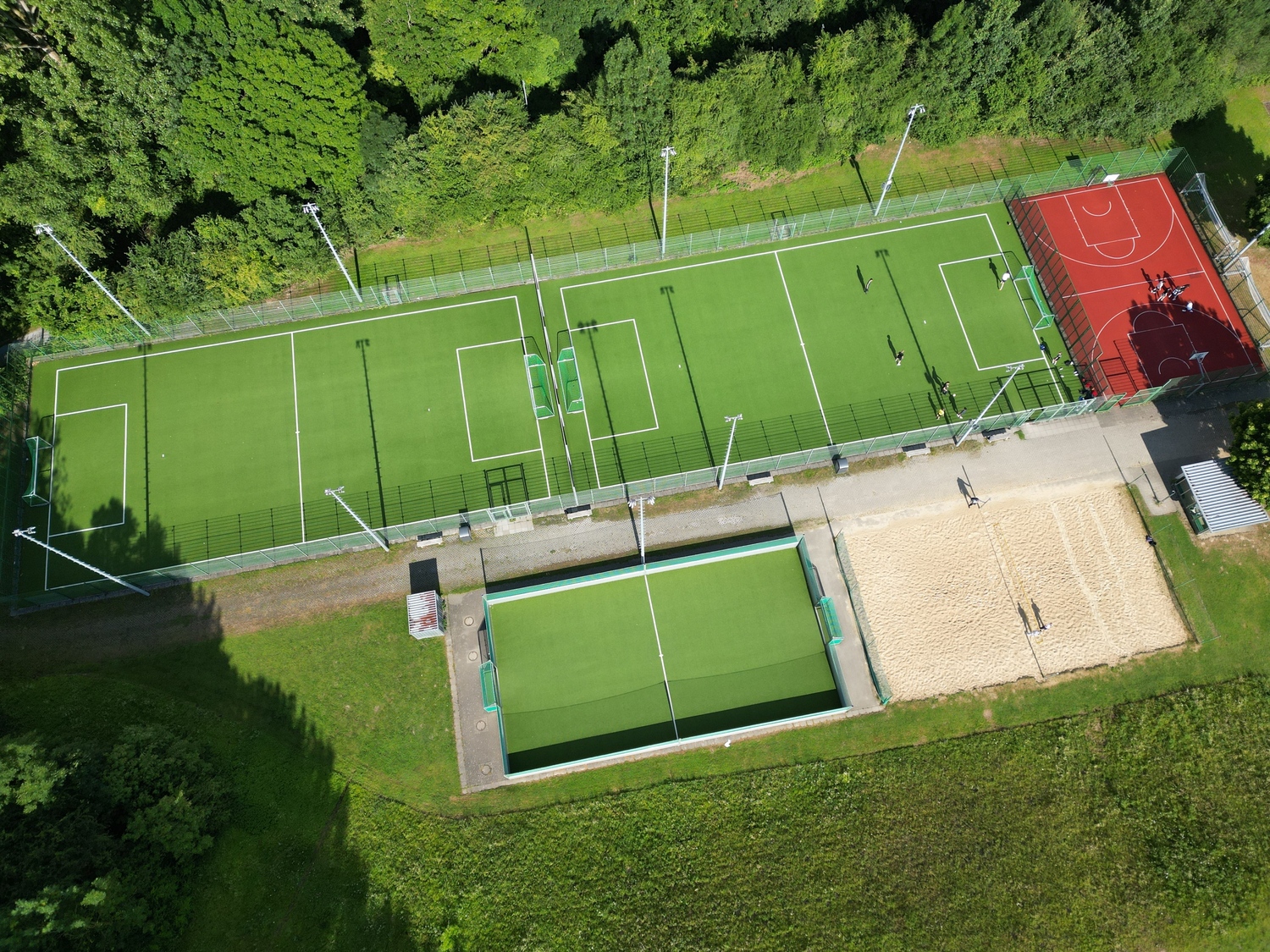 Luftaufnahmen des Kleinspielfelder-Bereichs auf dem Außengelände des Ahorn-Sportparks mit mehreren Sportflächen.  Zwei große Kunstrasenplätze liegen nebeneinander. Auf dem rechten großen Kunstrasenplatz spielen Personen. Von den großen Kunstrasenplätze ist rechts davon ist ein rotes Streetbasketballfeld mit Basketballkörben und spielenden Personen zu sehen. Unten rechts befindet sich ein Beachvolleyballfeld mit hellem Sand, auf dem ebenfalls Menschen spielen. Links der Beachvolleyballanlage befindet sich ein kleinerer Kunstrasenplatz. Die Anlage ist von Bäumen und Grünflächen umgeben.