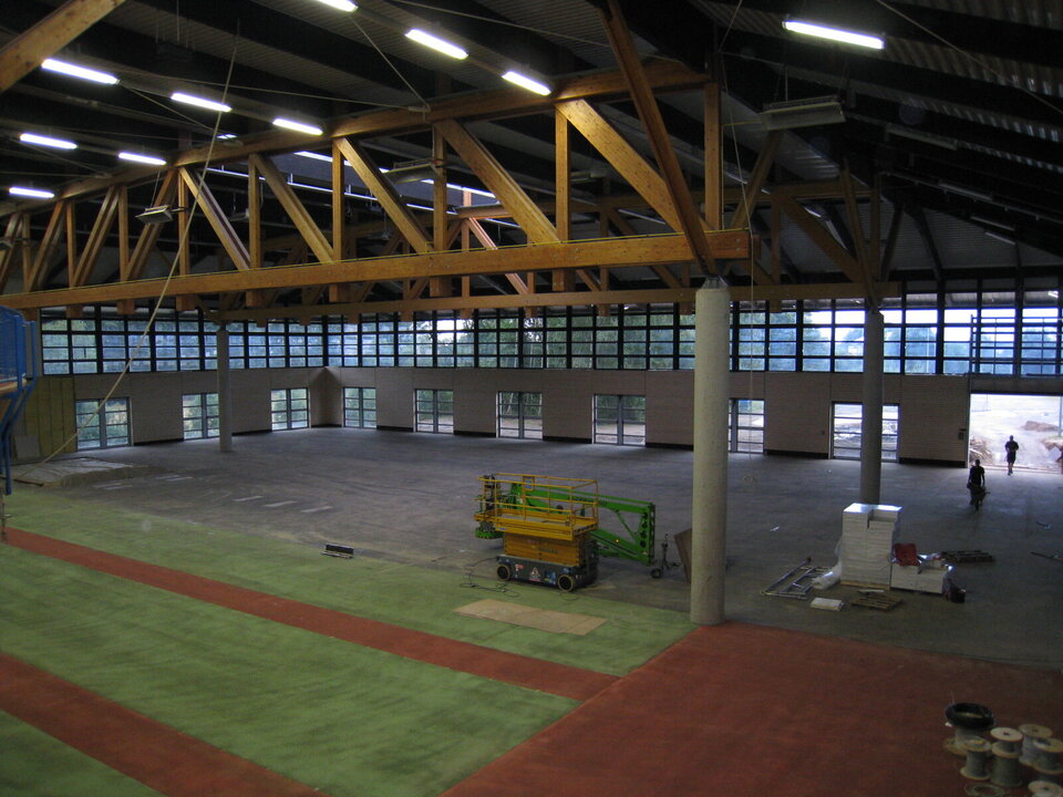 Dieses Foto zeigt einen Zwischenstand während des Bauabschnittes der Erweiterung der Ahorn-Sportpark-Halle an der Hallennordseite. Zu sehen ist, wie die Sporthalle bereits ab dem ersten Betonpfosten in der Mitte durch den offenen Anbau erweitert wurde. Im vorderen Bereich ist der Tatranboden in den Farben rot/grün abgebildet. Ab dem ersten Betonpfosten ist die Fläche schwarz und auf diesem stehen Baumaterialien. Zwei Menschen kommen über das offene Tor am rechten Rand des Bildes in den Bereich.