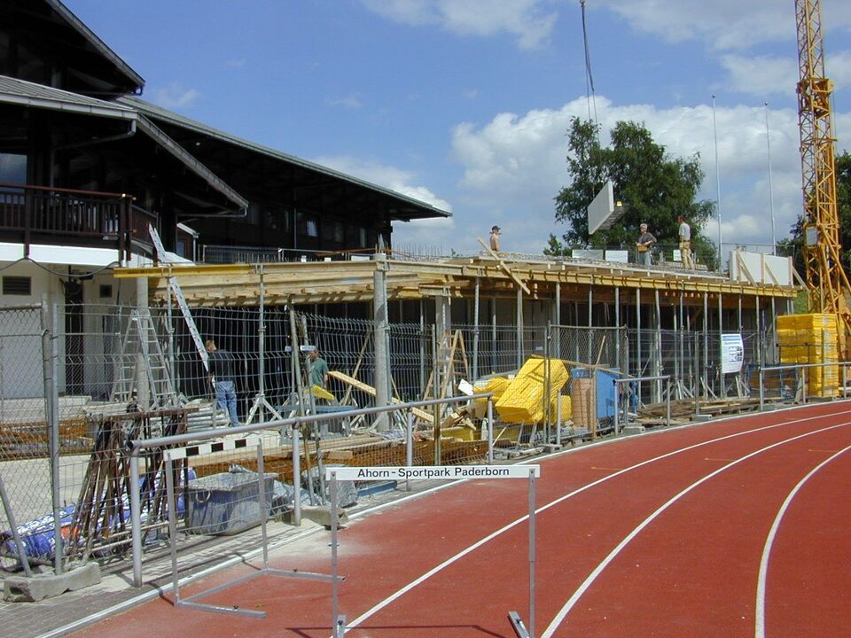 Angrenzent an der roten Tartanbahn des Leichtathletikstadion werden Stadionumkleiden gebaut. Der Rohbau aus einem Kontrukt aus Betonteilen, Metallpfleiler und Holsbretter ist vom einem grauen Bauzaun eingerahmt. Auf dem Rohbau sind Handwerker. Am rechten Rand steht ein gelber Baukran.