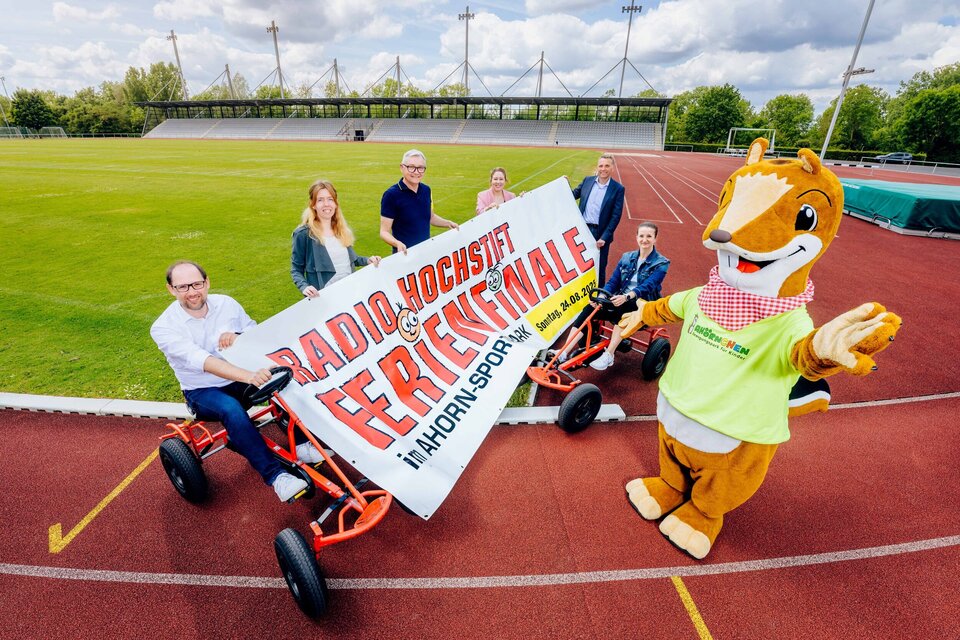 Im Leichtathletikstadion des Ahorn-Sportparks präsentieren von links nach rechts Ralf Pahlsmeier, Maren Richts, Christian Müller, Dania Tölle, Oliver Bekiersch, Jessica Spörhase und das Ahörnchen das Ankündigungsbanner für das Radio Hochstift-Ferienfinale. Dabei sitzen Ralf Pahlsmeier und Jessica Spörhase auf roten Kettcars. Auf dem Banner steht: Radio Hochstift-Ferienfinale im Ahorn-Sportpark 24.08.2025 11:00-18:00 Uhr.