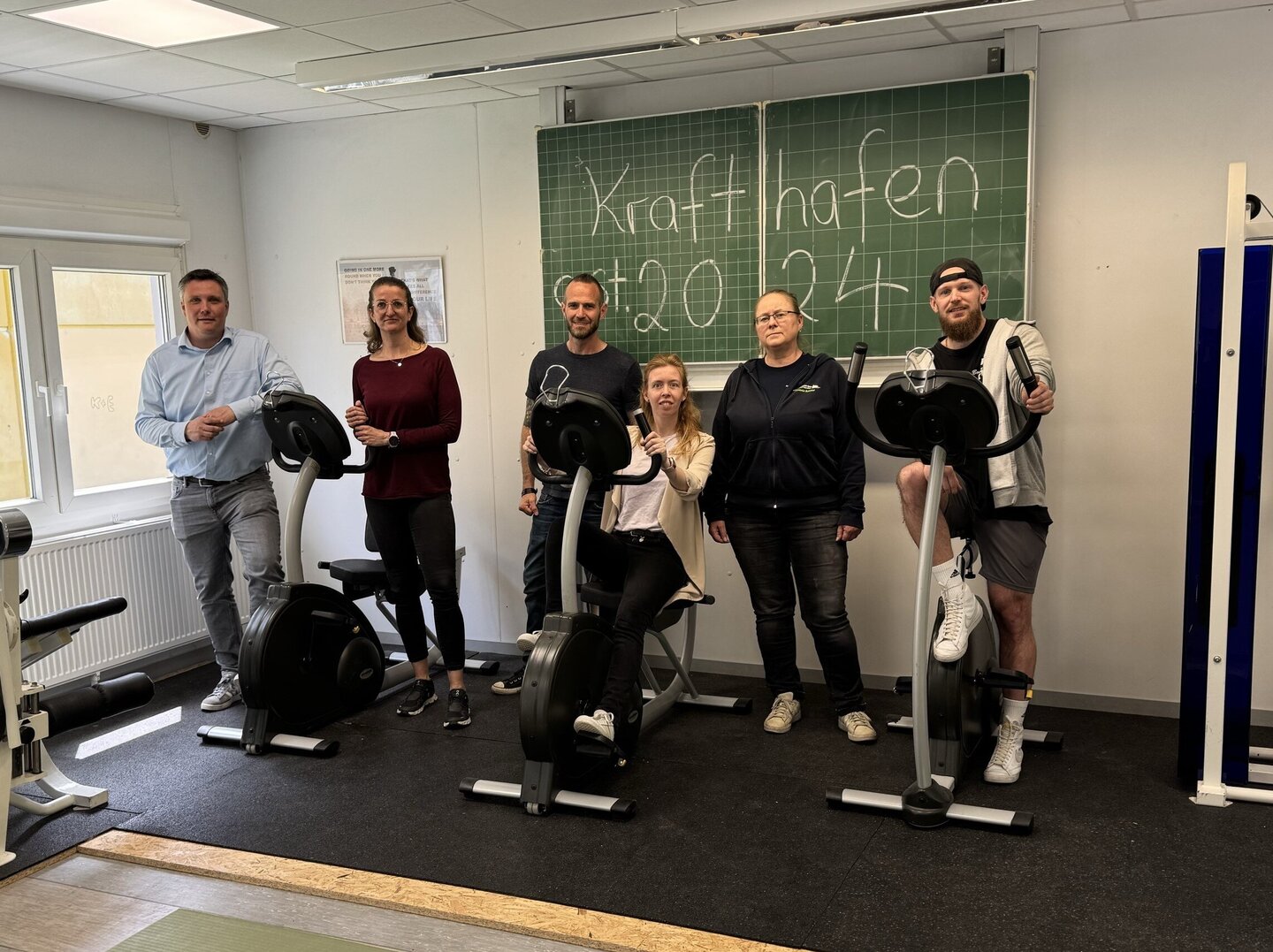 Im Krafthafen der Friedrich-Spee-Gesamtschule präsentieren Dr. Alexander Hennemann, Jessica Spörhase, Dr. Daniel Raths, Maren Richts, Stefanie Baldauf und Jochen Rehkämper drei Cardiogeräte. Die Cardiogeräte stehen auf einem Podest, welcher mit einem schwarzen Belag belegt ist. Im Hintergrund ist eine Schultafel aufgehangen auf welcher Krafthafen 2024 steht.