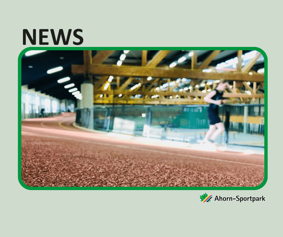 Das Bild zeigt die 200-Meter-Rundlaufbahn im Ahorn-Sportpark. Im Vordergrund ist der rötlich-gummierte Boden der Bahn zu sehen. Eine einzelne Person joggt entlang der Bahn und ist leicht unscharf dargestellt, was die Bewegung betont. Im Hintergrund erkennt man die große, helle Sporthalle mit sichtbaren Holzträgern an der Decke und einer modernen Beleuchtung. Die Bahn ist von einem Sicherheitsnetz und Glaswänden umgeben. In der oberen linken Ecke steht in großer Schrift „NEWS“, unten rechts ist das Logo des Ahorn-Sportparks abgebildet.