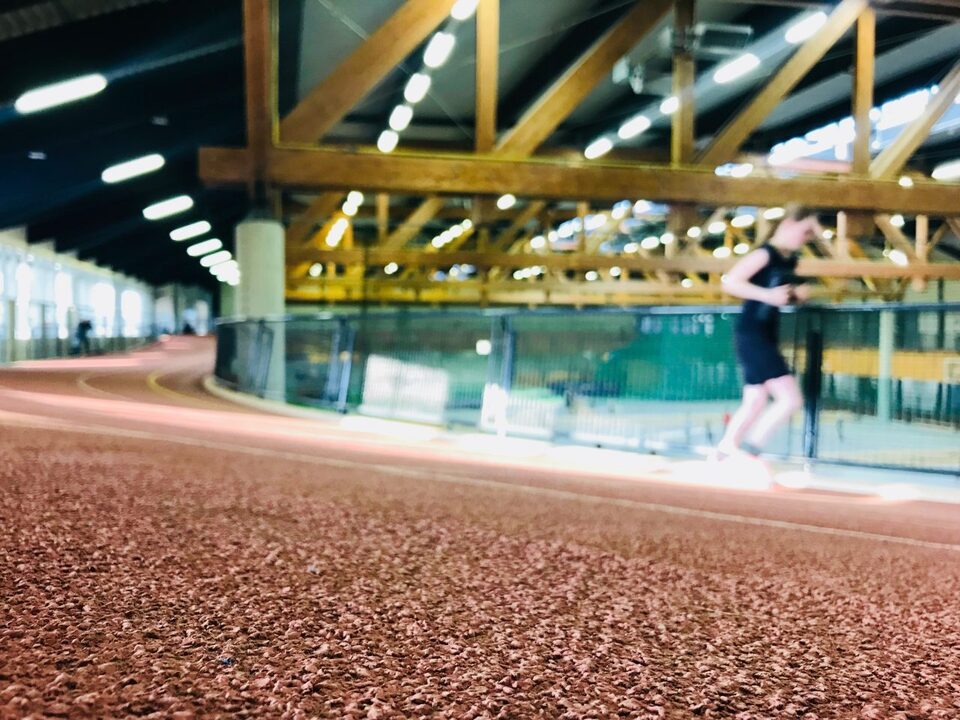 Das Bild zeigt die 200-Meter-Rundlaufbahn im Ahorn-Sportpark. Im Vordergrund ist der rötlich-gummierte Boden der Bahn zu sehen. Eine einzelne Person joggt entlang der Bahn und ist leicht unscharf dargestellt, was die Bewegung betont. Im Hintergrund erkennt man die große, helle Sporthalle mit sichtbaren Holzträgern an der Decke und einer modernen Beleuchtung.