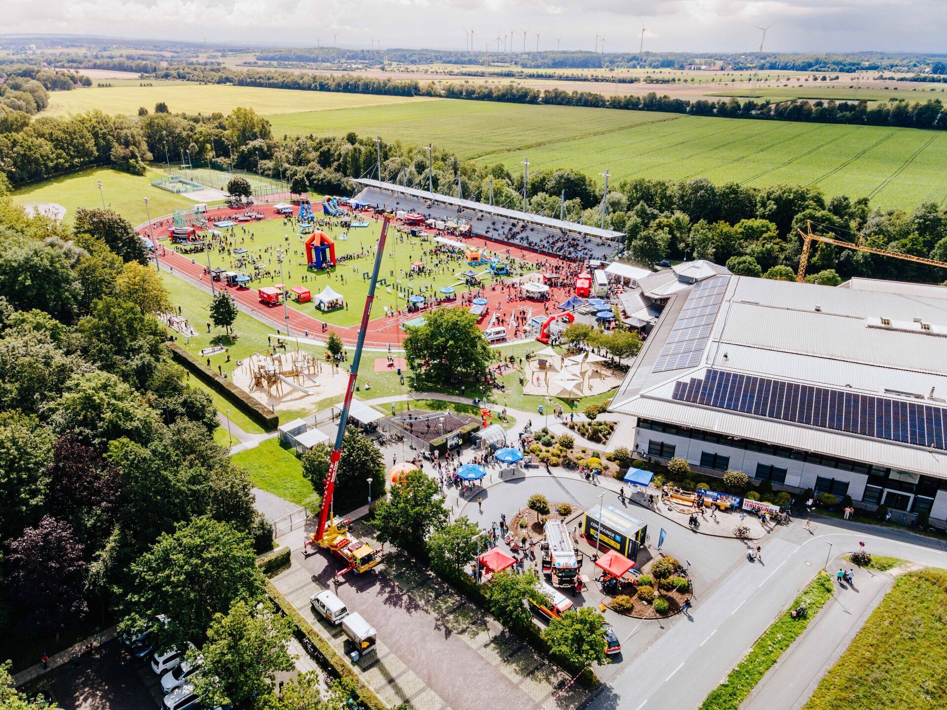 Die Luftaufnahme zeigt das Radio Hochstift-Ferienfinale im Ahorn-Sportpark. Im vorderen Bereich sind verschieden Stände wie z. B. mit einem Feuerwehrauto oder einem Ballon am Kran zu sehen. Im hinteren Teil befindet sich das Leichathletikstadion, wo mehrere bunte Hüpfburgen, Spielstationen und Pavillons aufgebaut sind. Zahlreiche Menschen sind in den beiden Bereichen unterwegs. Rechts neben dem Stadion steht ein großes Gebäude mit Solarpanelen auf dem Dach.
