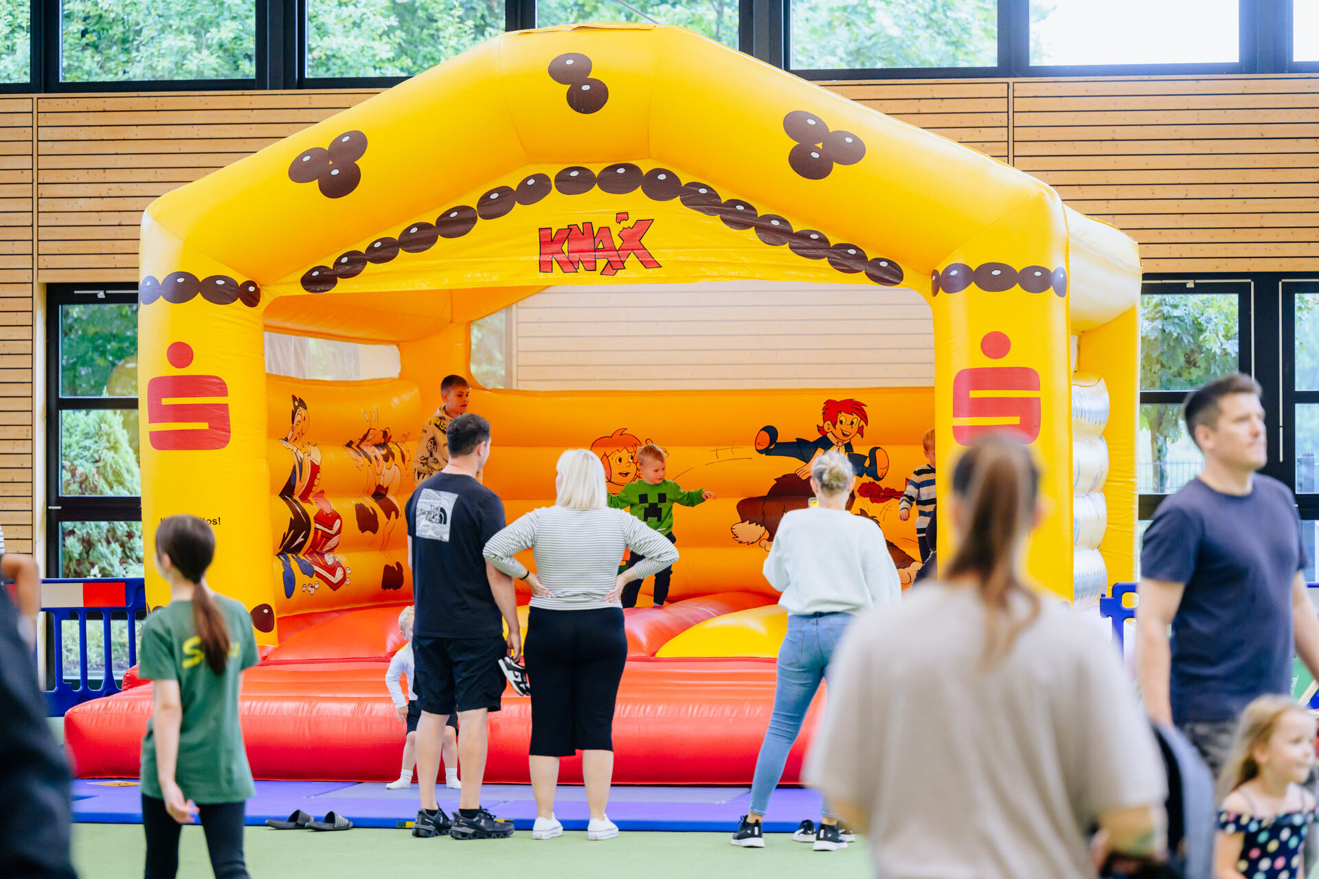 Vor einer gelb-roten Hüpfburg mit dem Sparkassen-Logo und den worten "KNAX" stehen mehrere Menschen. Auf der Hüpfburg hüpfen mehrere Kinder.