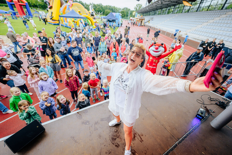 Isa Glücklich steht steht auf der Bühne beim Radio Hochstift-Ferienfinale und macht ein Selfie mit ausgestreckten Armen. Im Hintergrund jubeln zahlreiche Kinder und Erwachsene, die dicht gedrängt vor der Bühne stehen. Alle wirken gut gelaunt und freuen sich. Links im Bild sieht man viele lachende Kinder, einige winken oder rufen. Rechts im Hintergrund steht das Maskottchen Luzi mit ausgebreiteten Armen. Im Stadionbereich sieht man Tribünen mit leeren Sitzreihen und auf dem Rasen im Hintergrund sind bunte Hüpfburgen und weitere Besucher zu erkennen