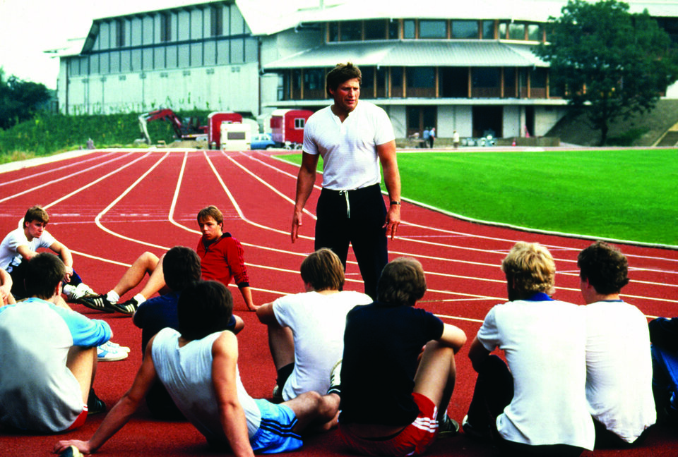 Im Leichtathletikstadion des Ahorn-Sportparks steht Kurt Bendlin vor sitzenden Menschen. Im Hinterhrund ist das Gebäude des Ahorn-Sportparjs zu sehen und davor sind Bauarbeiten.