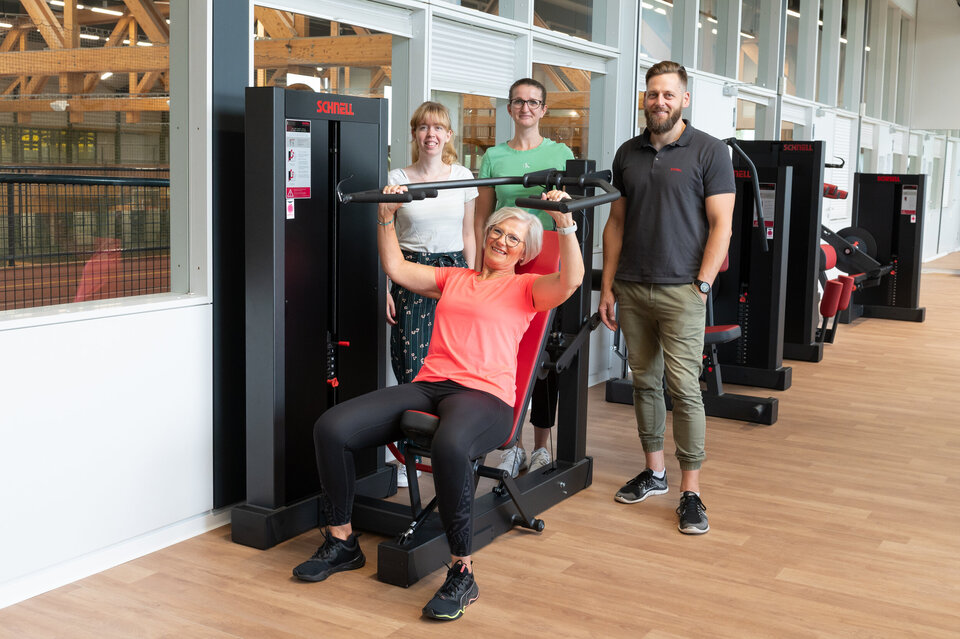 Der Ausschnitt zeigt einen Teil des neu gestalteten Fitnessraumes im Ahorn-Sportpark. Mehrere schwarz-Rote Fitnessgeräte sind zu sehen. An dem Fitnessgerät im Vordergrund stehen drei Personen hinter dem Gerät und eine Person sitzt auf dem Sitz des Fitnessgerätes und zieht mit den Armen die Armhalterung heruntern.