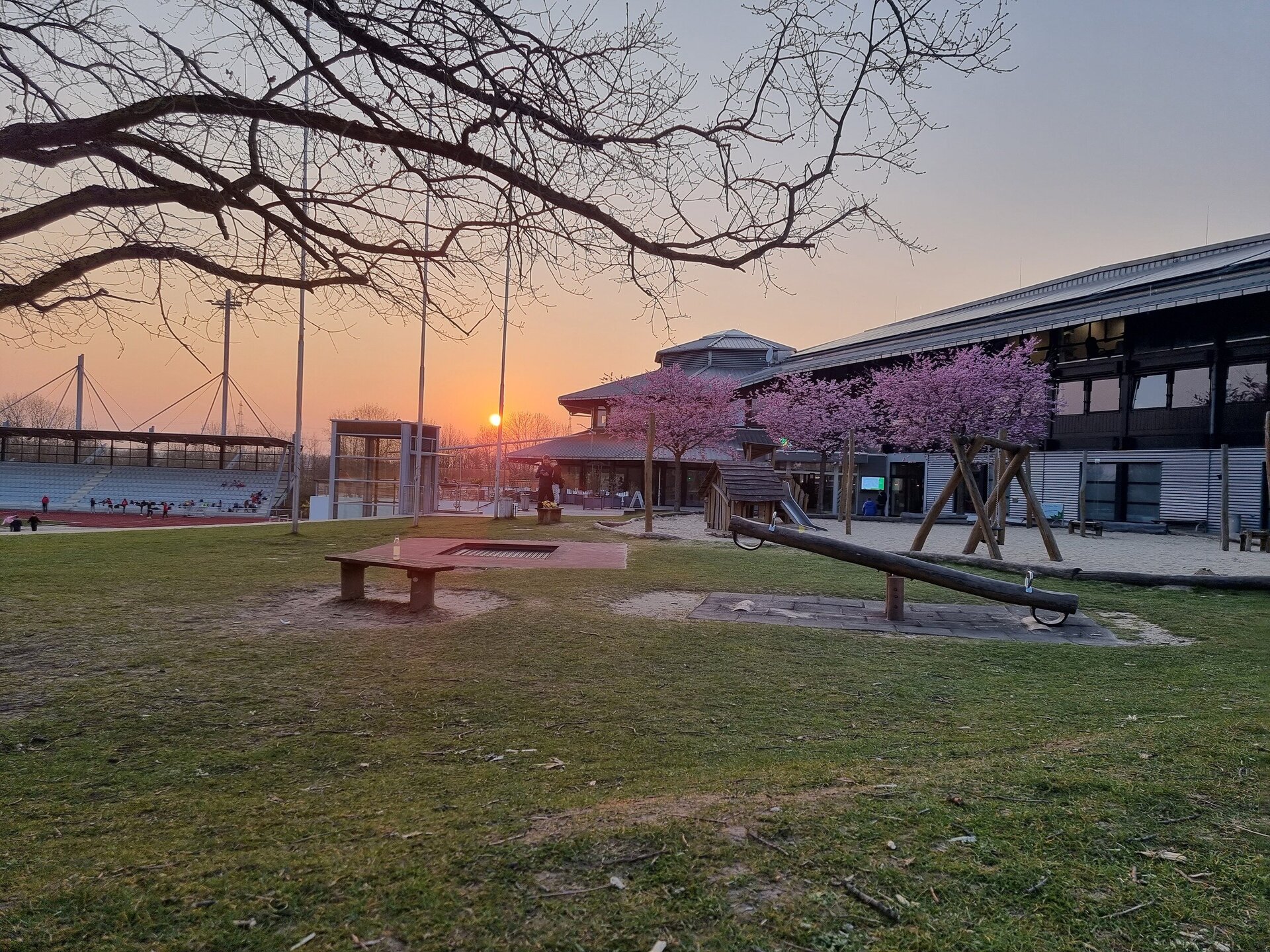 Im Vordergrund steht der Ahörnchen-Spielplatz des Ahorn-Sportparkes mit einer Holz-Wippe, einer Schaukel, eine Holzhütte, eine kleine Rutsche, einer Bank und blühenden Kirschbäumen. Im Hintergrund befindet sich die Ahorn-Sportpark-Halle auf der rechten Seite und auf der linken Seite das Leichtathletikstadion mit einer Tribüne. Im Hintergrund geht die Sonne unter.