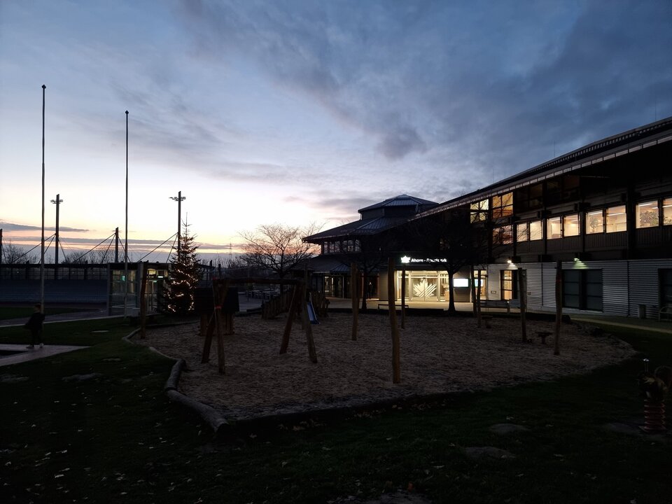Der Ahörnchen-Spielplatz des Ahorn-Sportparks mit Holzgeräten auf Sandfläche bei Dämmerung und links daneben und ein Weihnachtsbaum mit Lichtern steht im Vordergrund. Im Hintergrund ist das Gebäude des Ahorn-Sportparks mit dem beleuchteten Eingang zu sehen.