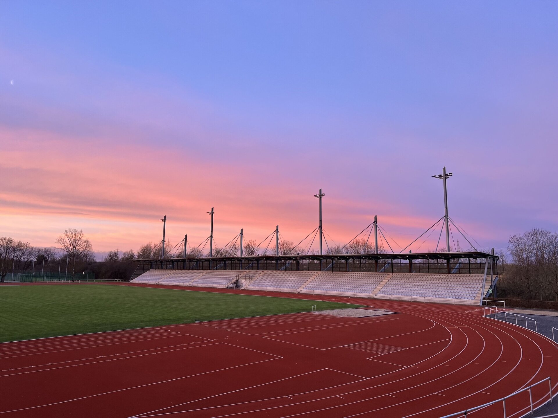 Das leere Leichtathletikstadion mit roter Laufbahn und Tribüne, umgeben von Bäumen. Der Himmel ist in sanften Rosa-, Lila- und Blautönen gefärbt, während eines Sonnenuntergangs.