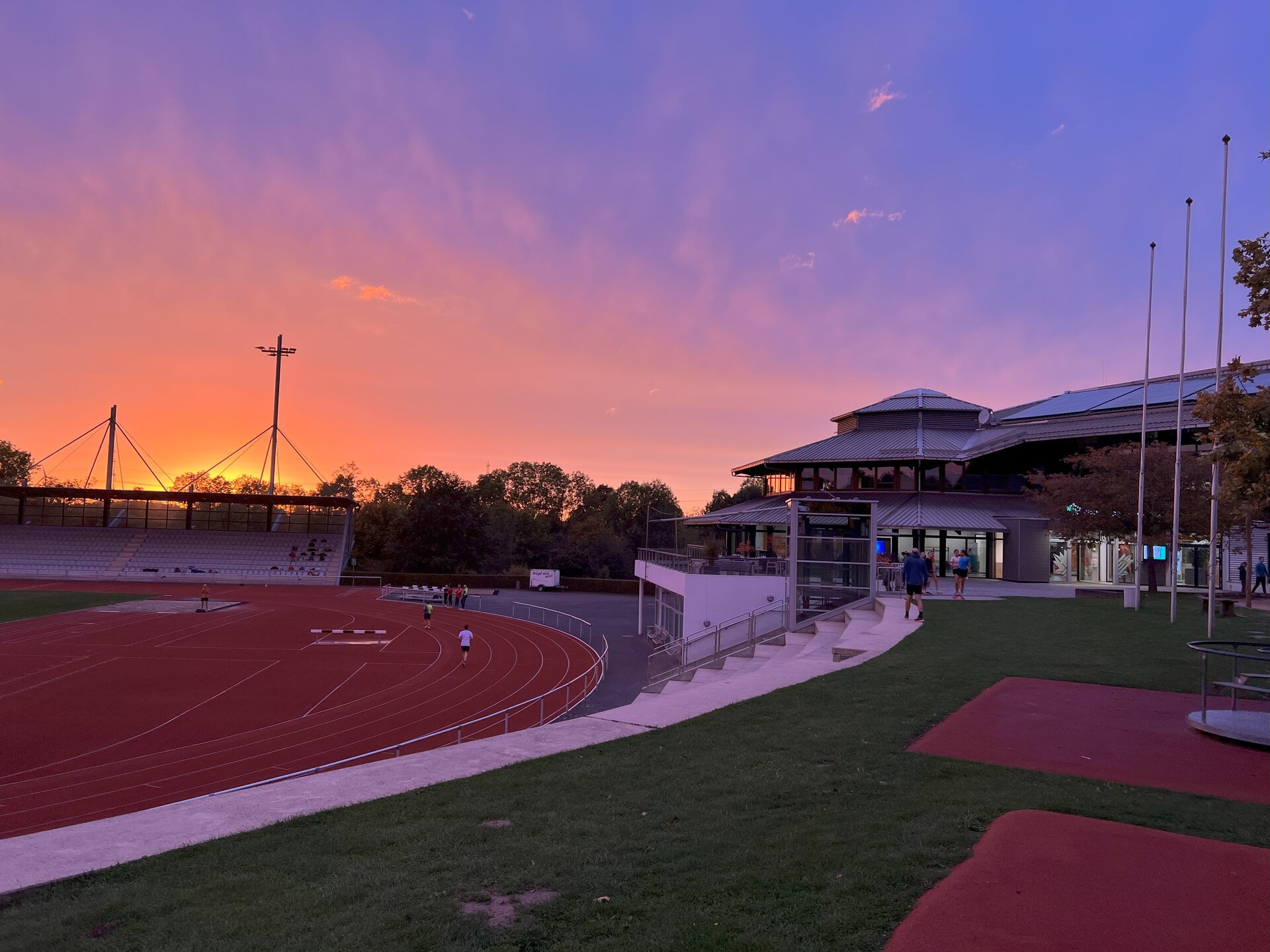 Der Himmel über dem Leichtathletikstadion und der Ahorn-Sportpark-Halle ist in sanften Rosa-, Lila- und Blautönen während eines Sonnenunterganges gefärbt. Hinter der Tribüne geht die Sonne unter. Auf der linken Seite des Bildes ist das Stadion. Dort sind Sportlinnen und Sportler aktiv. Auf der rechten Seite ist der Eingangsbereich der Ahorn-Sportpark-Halle, wo sich Menschen aufhalten.