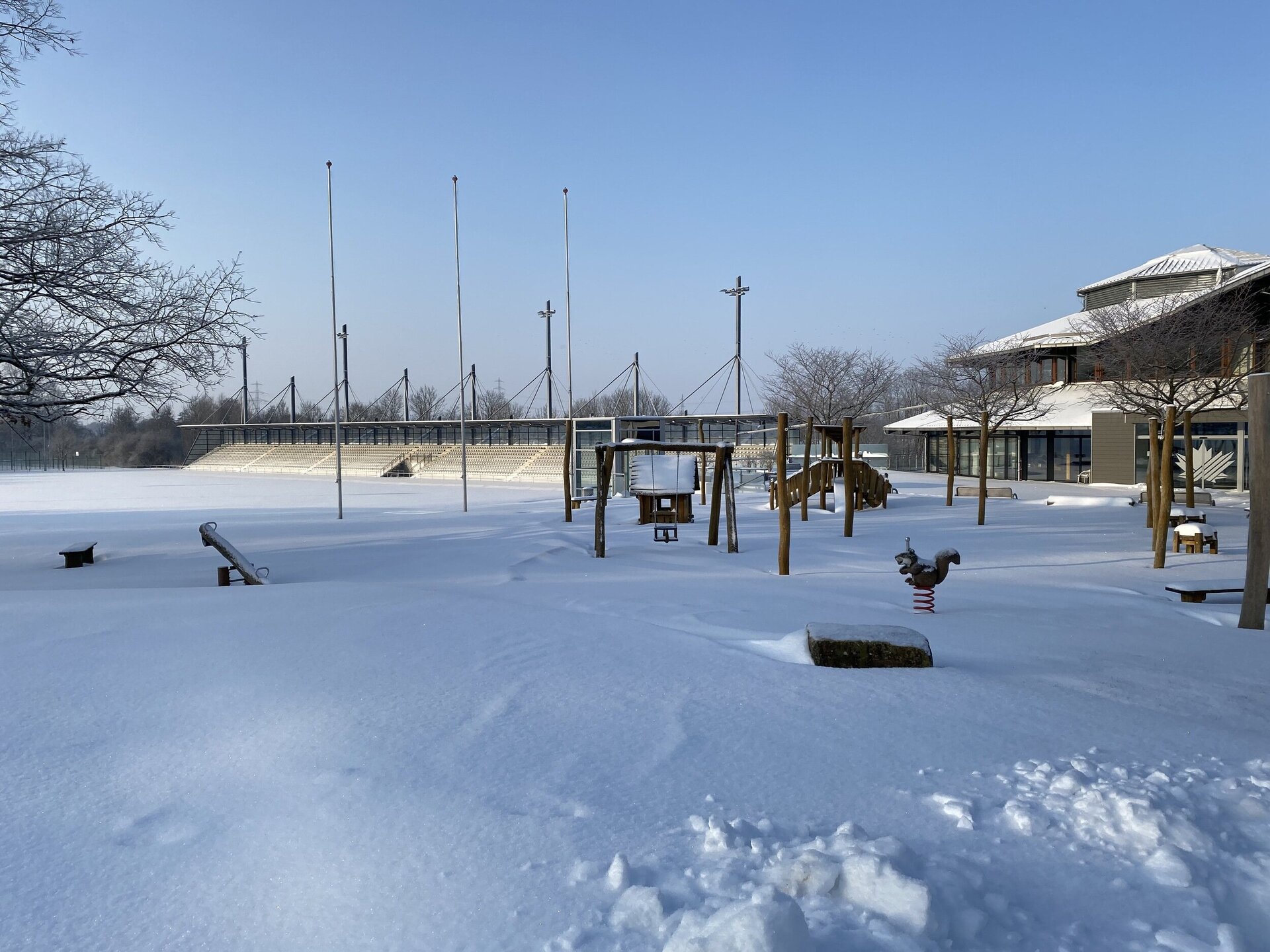 Der Boden ist mit Schnee bedeckt. Die Spielgeräte des Ahörnchen-Spielplatzes stehen inmitten der Schneedecke. Auch das Leichtathletikstadion ist voller Schnee, und auf dem Dach der angrenzenden Halle, welches im rechten Hintergrund zu sehen ist, liegt ebenfalls Schnee. Am Himmel ist keine Wolke zu sehen, er ist klar und blau.
