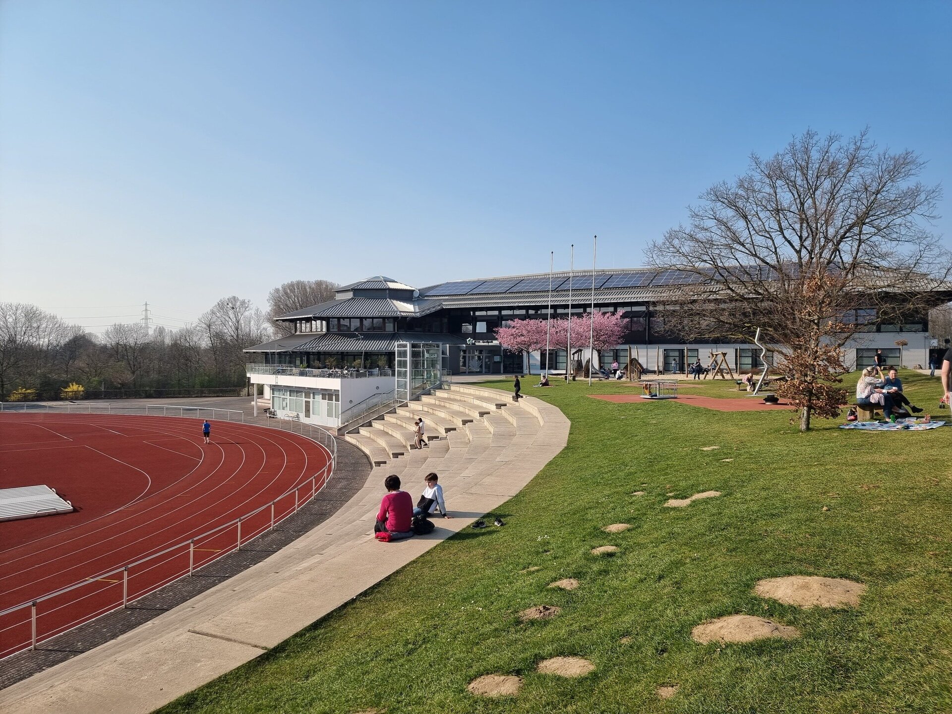 Auf dem Ahörnchen-Spielplatz und der angrenzenden Betontribüne halten sich mehrere Menschen auf. Links im Bild ist das Leichtathletikstadion zu sehen, in dem eine Person läuft. Im Hintergrund stehen drei rosa blühende Kirschbäume und die Ahorn-Sportpark-Halle.