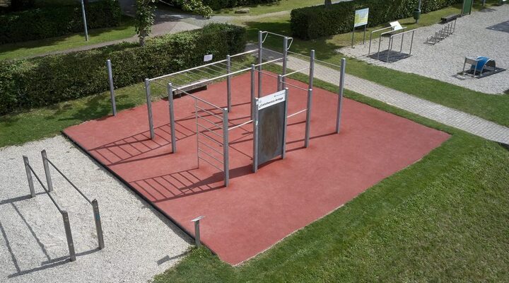 Vogelperspektive auf die Calisthenics-Anlage mit rotem Fallschutzboden. Links daneben steht ein Metallbarren auf Schotteruntergrund. Rechts verläuft ein Fußweg, an dem ein weiterer Bereich mit mehrere Outdoor-Fitnessgeräten mit einem Schotteruntergrund beginnt.