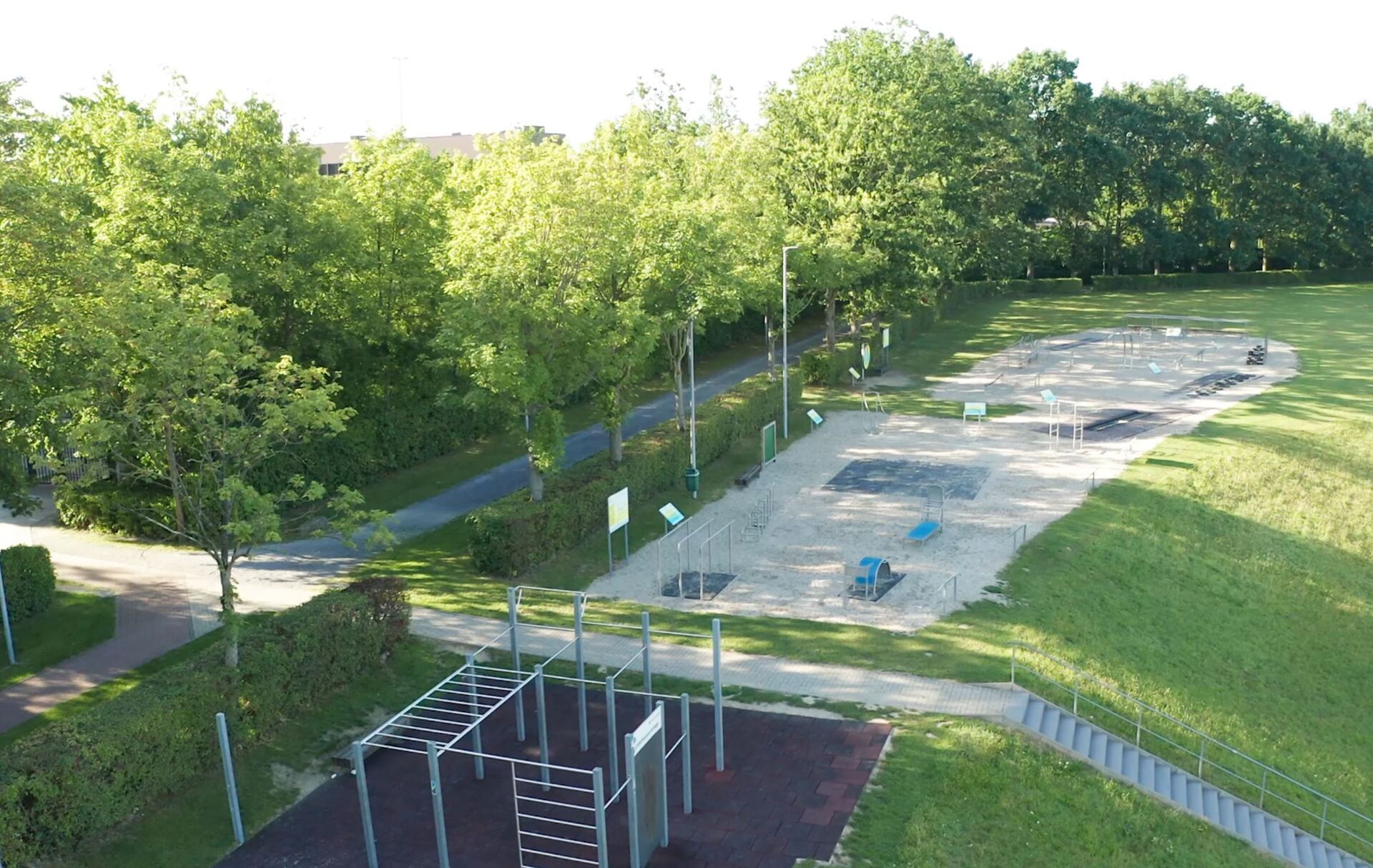 Im unteren Bereich des Bildes befindet sich die Calisthenics-Anlage und im oberen Bereich den 4°Circle des Outdoor-Fitnessparcours. Zwischen den beiden Bereiche verläuft ein ein Fußweg mit einer Treppe. Links im Bild stehen merere grüne Bäume und eine lange grüne Hecke.