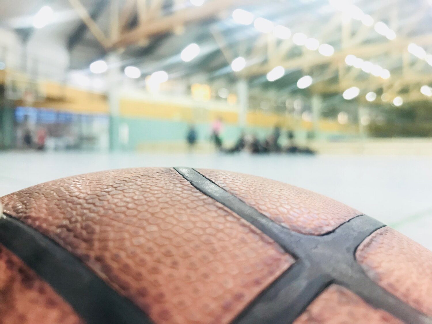 Im Vordergrund ist ein Basketball zur Hälfte sichtbar. Im Hintergrund sind mehrere Ballspielflächen zu erkennen, auf denen sich Personen aufhalten. Der Hintergrund ist unscharf dargestellt.
