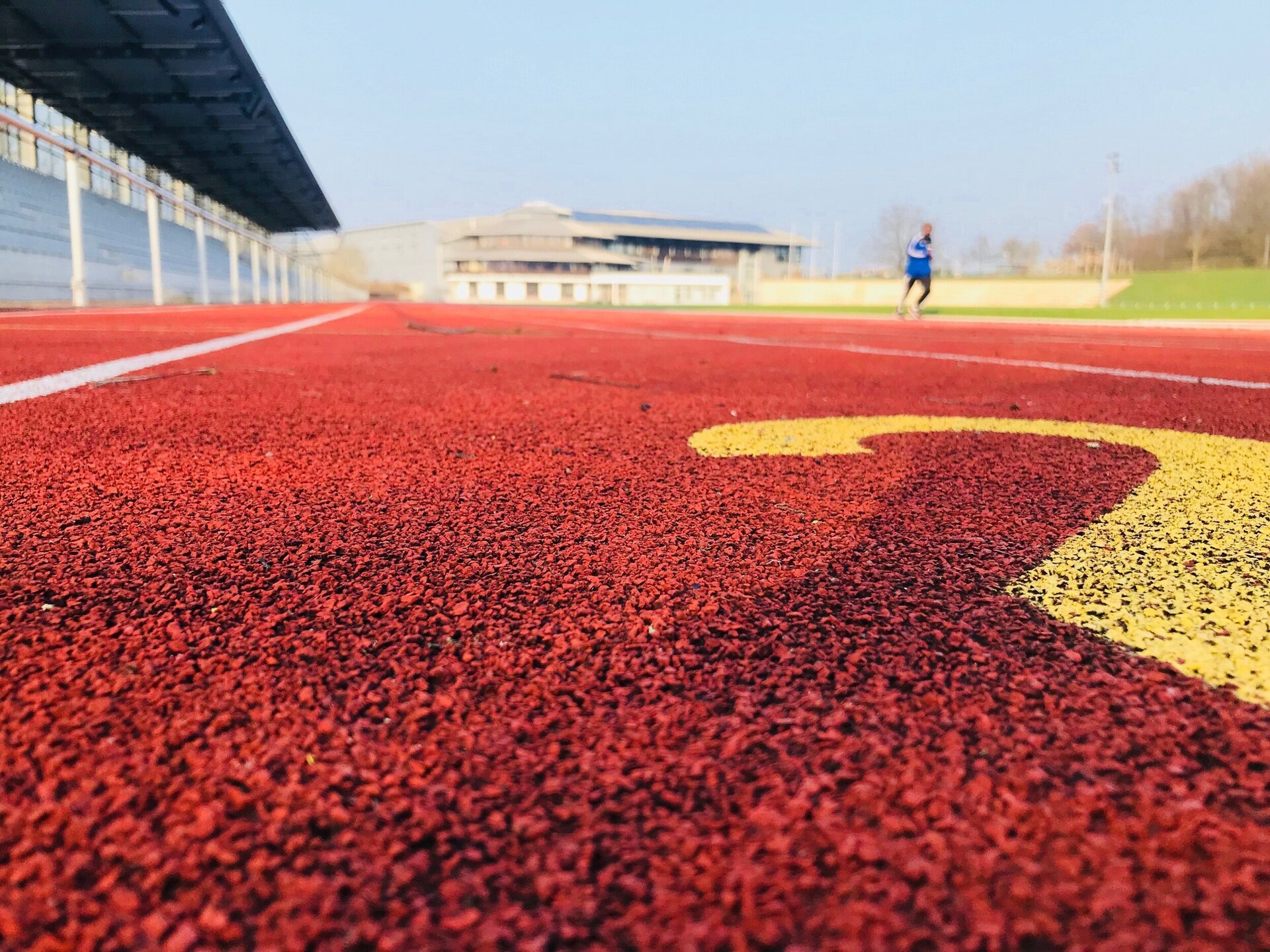 Das Leichtathletikstadion mit roter Tartanbahn. Auf der Bahn läuft eine Person. Links im Bild ist eine Zuschauertribüne zu sehen und im Hintergrund steht eine Ahorn-Sportpark-Halle.