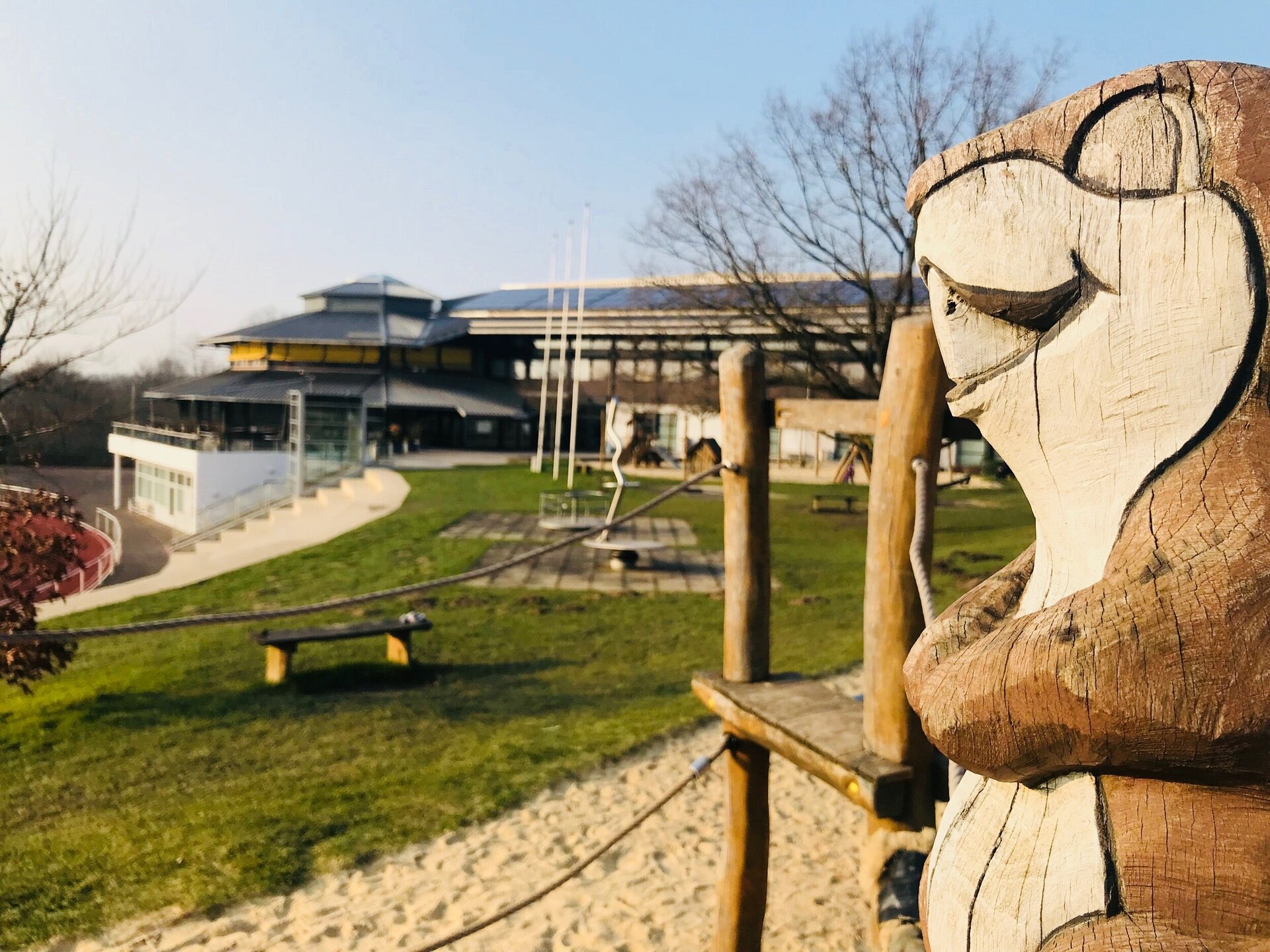 Mehrere Spielgeräte des Ahörnchen-Spielplatzes sind im Vordergrund zu sehen. Rechts am Bildrand steht als Holzfigur das Ahörnchen. Im Hintergrund ist die Ahorn-SPortpark-Halle zu sehen.
