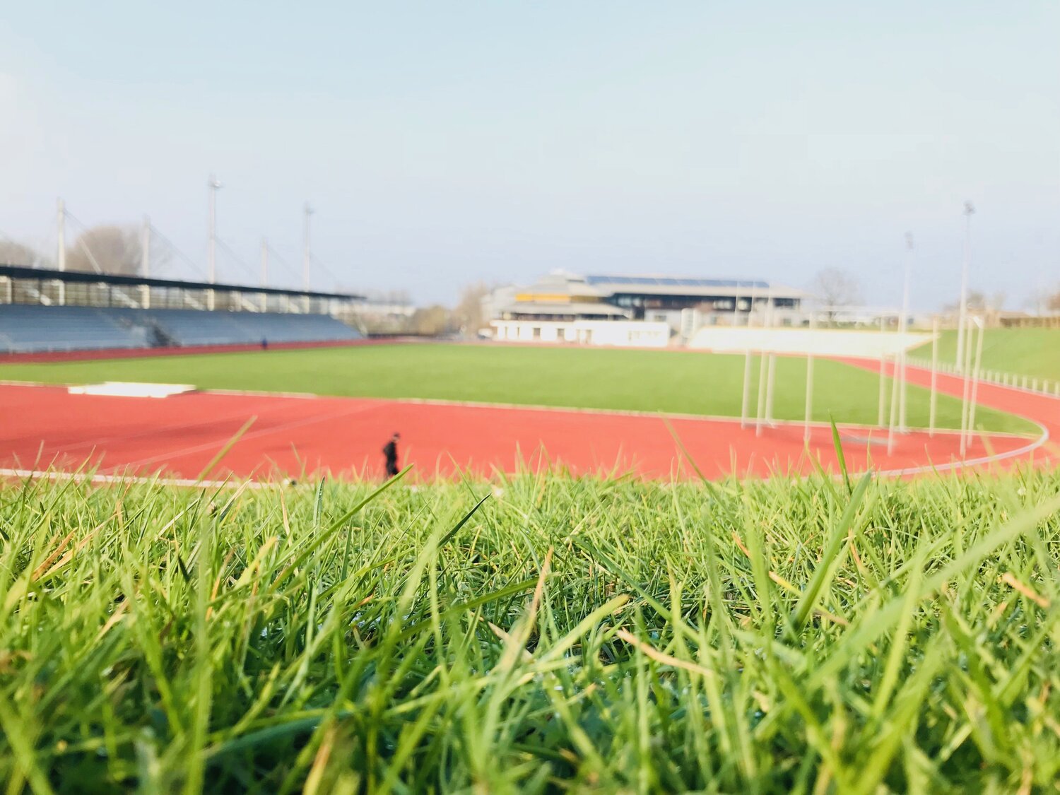 Das Leichtathletikstadion ist im Vordergrund zu sehen. Dort läuft ein Läufer. Im Stadion sind keine Leichtathletikaufbauten vorhanden, sondern nur die Metallrahmen für die Module. Links grenzt die Tribüne an und weit im Hintergrund ist die Ahorn-Sportpark-Halle zu sehen.