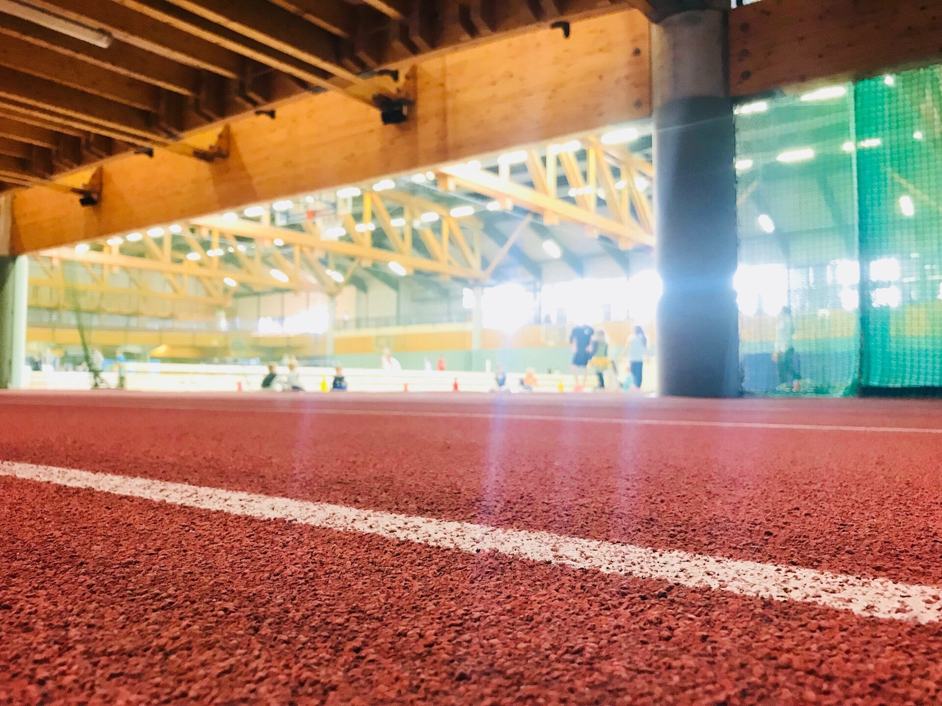 Sprintbahn in einer Sporthalle mit weißen Linien im Vordergrund. Dahinter befinden sich Ballspielflächen, auf denen Menschen spielen. Die Ballspielflächen sind unscharf dargestellt.
