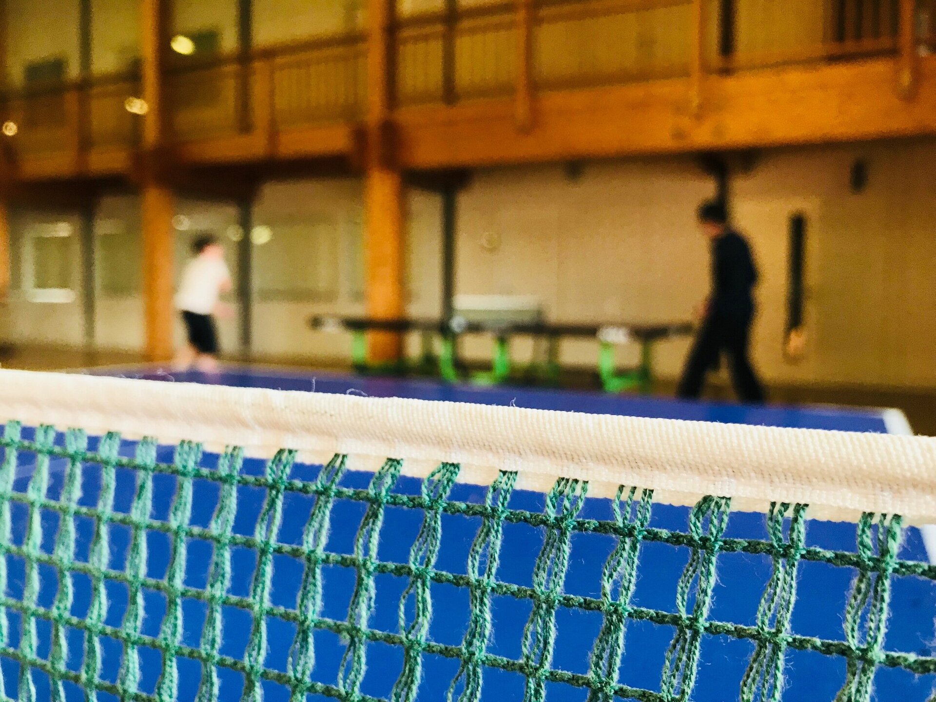 Nahaufnahme eines Tischtennisnetzes einer Tischtennisplatte im Vordergrund. An einer zweiten Tischtennisplatte im unscharfen Hintergrund spielen zwei Spieler.