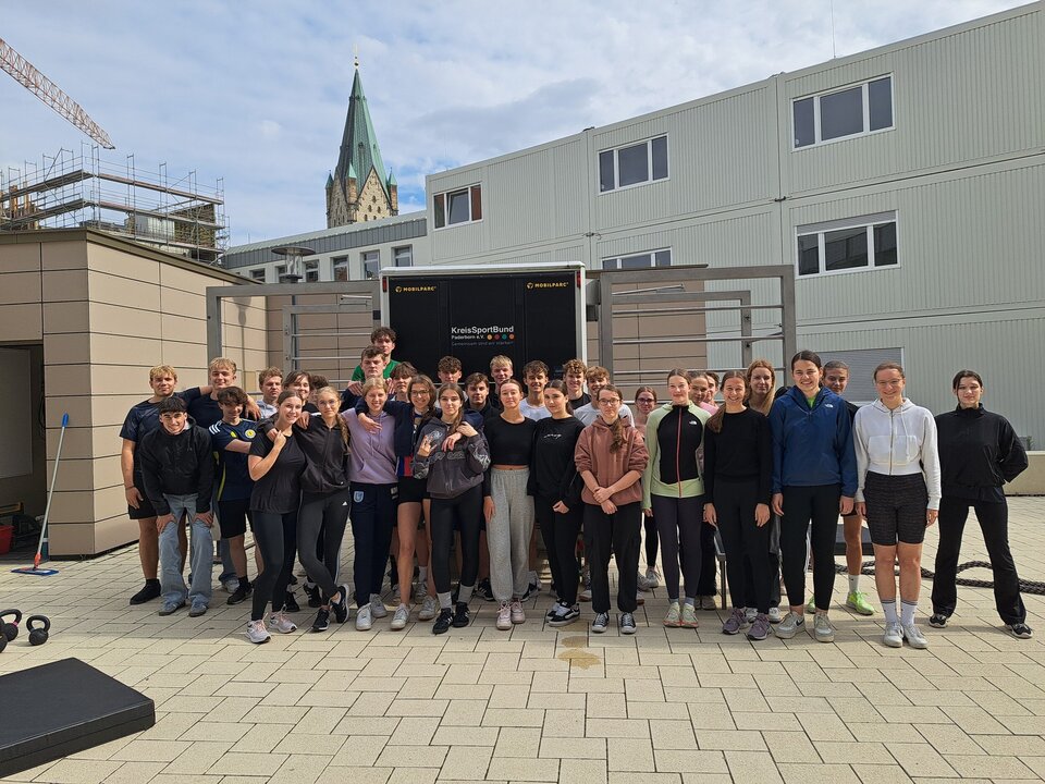 Schüler und Schülerinnen des Gymnasiums St. Michael stehen auf dem schulhof vor dem Calisthenics-Anhänger. Im Hintergrund befindet sich ein Schulgebäude und die Turmspitze des Paderborner Doms ist  oberhalb des Gebäudedaches zu sehen.