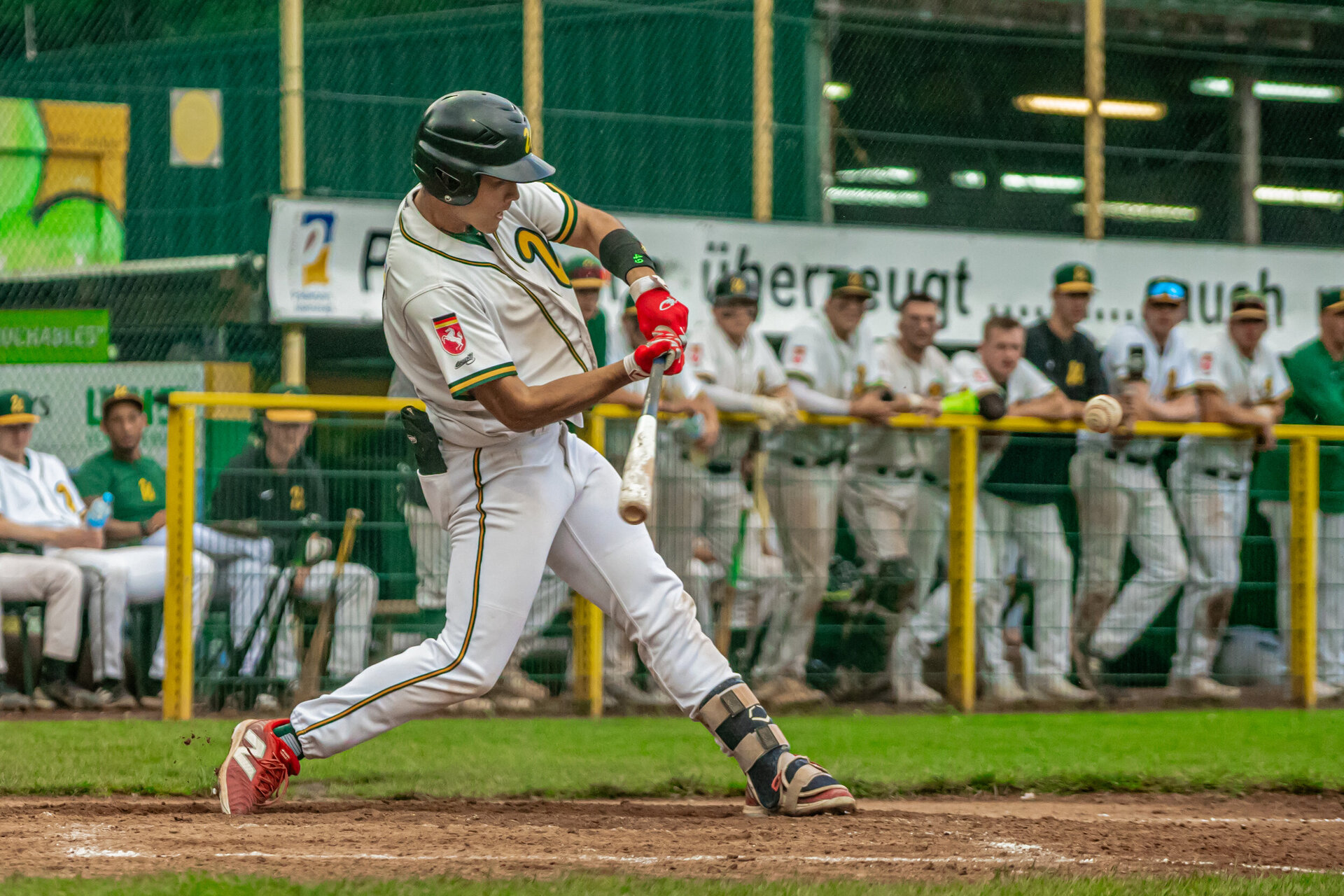 Ein Baseballspieler von den Untouchables Paderborn, in weißer Uniform mit gelben und grünen Streifen, trifft mit dem Schläger den Ball. Er trägt einen schwarzen Helm, rote Handschuhe und Beinschützer. Im Hintergrund stehen Teamkollegen in weiß-grünen Uniformen hinter einem gelben Zaun und beobachten das Spiel.