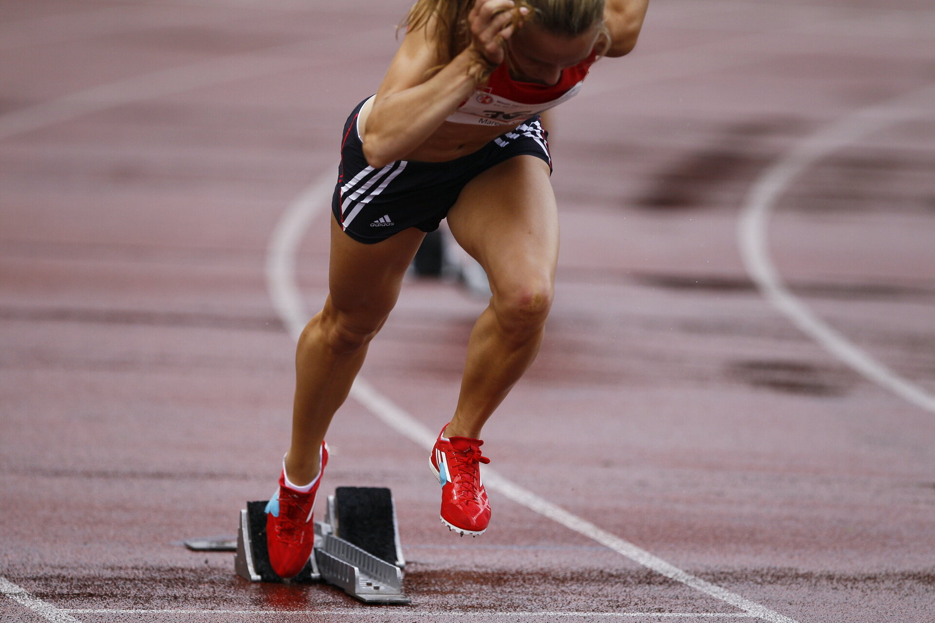 Eine Leichtathletin stößt sich kraftvoll aus dem Startblock ab. Sie trägt rote Spikes, schwarze kurze Sporthosen mit weißen Streifen und ein rotes Oberteil. Die Laufbahn ist nass, was auf Regen hindeutet