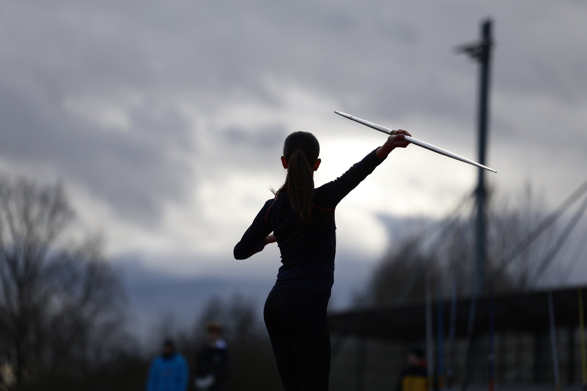 Eine Athletin steht im Leichtathletikstadion und bereitet sich auf den Speerwurf vor. Sie hält den Speer mit ausgestrecktem Arm über der Schulter, während der Himmel im Hintergrund bewölkt ist.