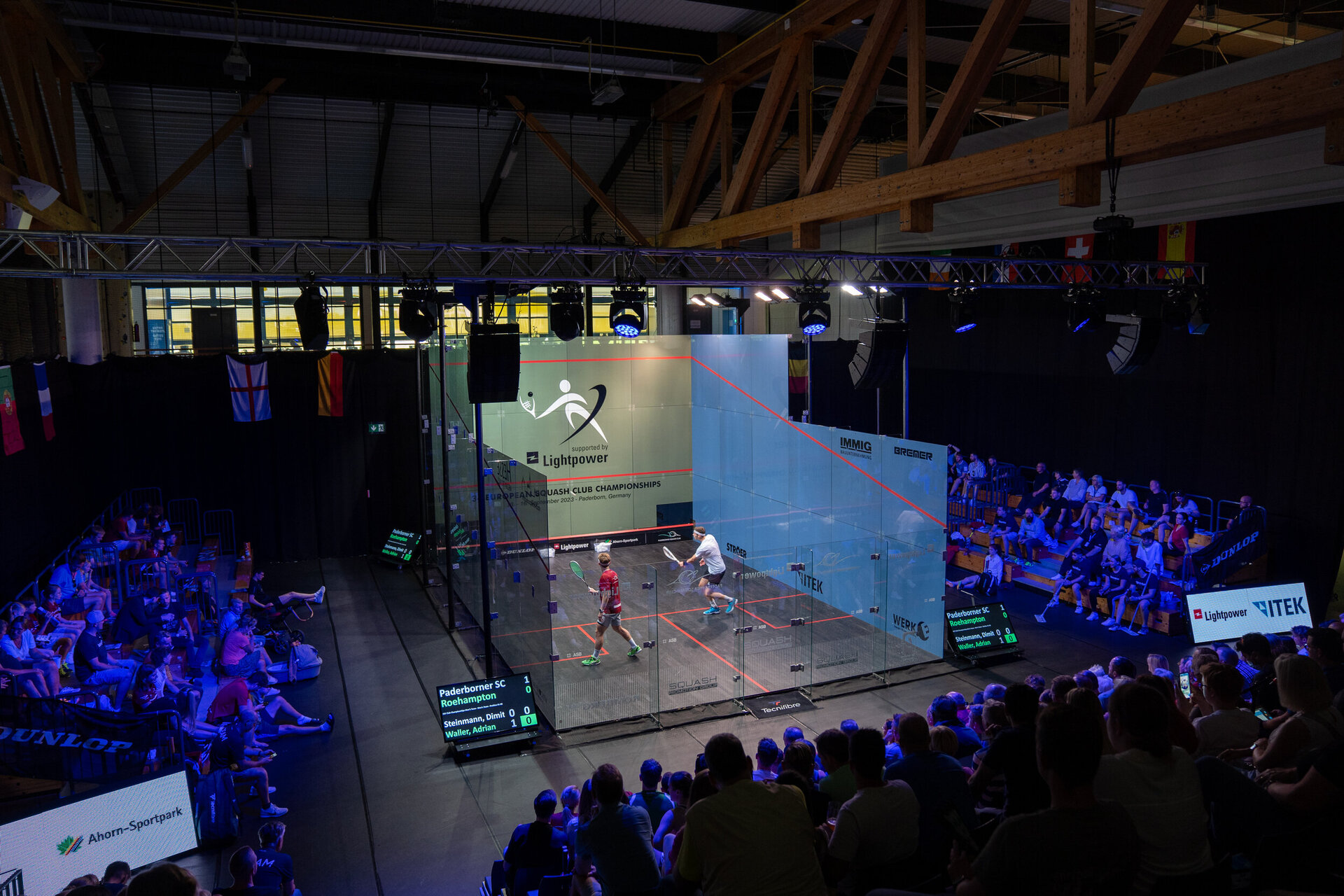 Blick in den beleuchtetes Glac-Court während des ECC 2023. Zwei Spieler befinden sich im Glas-Court, einer in rotem Trikot, der andere in weißem. Zuschauer sitzen auf beiden Seiten der Arena, während Anzeigetafeln Spielstände und Spielernamen anzeigen. Über dem Spielfeld hängen Flaggen und Scheinwerfer.