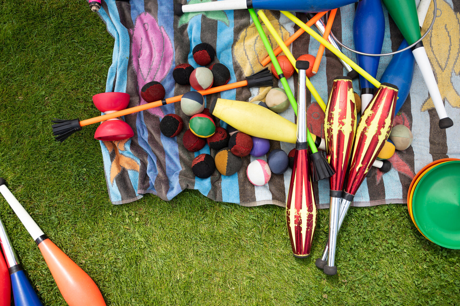 Verschiedenes Jonglier- und Zirkusmaterial liegt auf einer bunten Decke auf einer Wiese. Zu sehen sind Jonglierkeulen, Bälle, Flowersticks, Diabolos und bunte Jonglierteller.