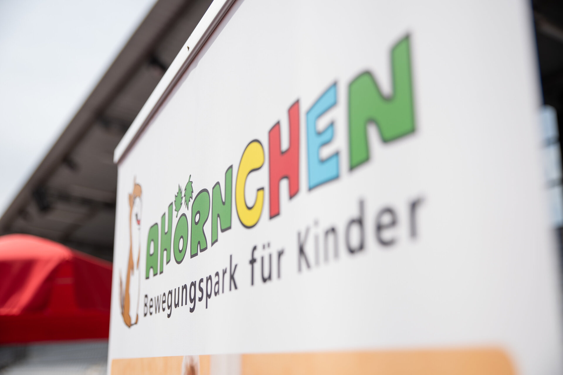 Im Vordergrund steht ein Schild mit der Aufschrift „AHÖRNCHEN – Bewegungspark für Kinder“ und links neben dem Text ist ein gezeichnetem Ahörnchen abgebildet. Die Buchstaben vom Ahörnchen sind bunt. Im Hintergrund ist ein roter Sonnenschirm zu erkennen und ein Dach.