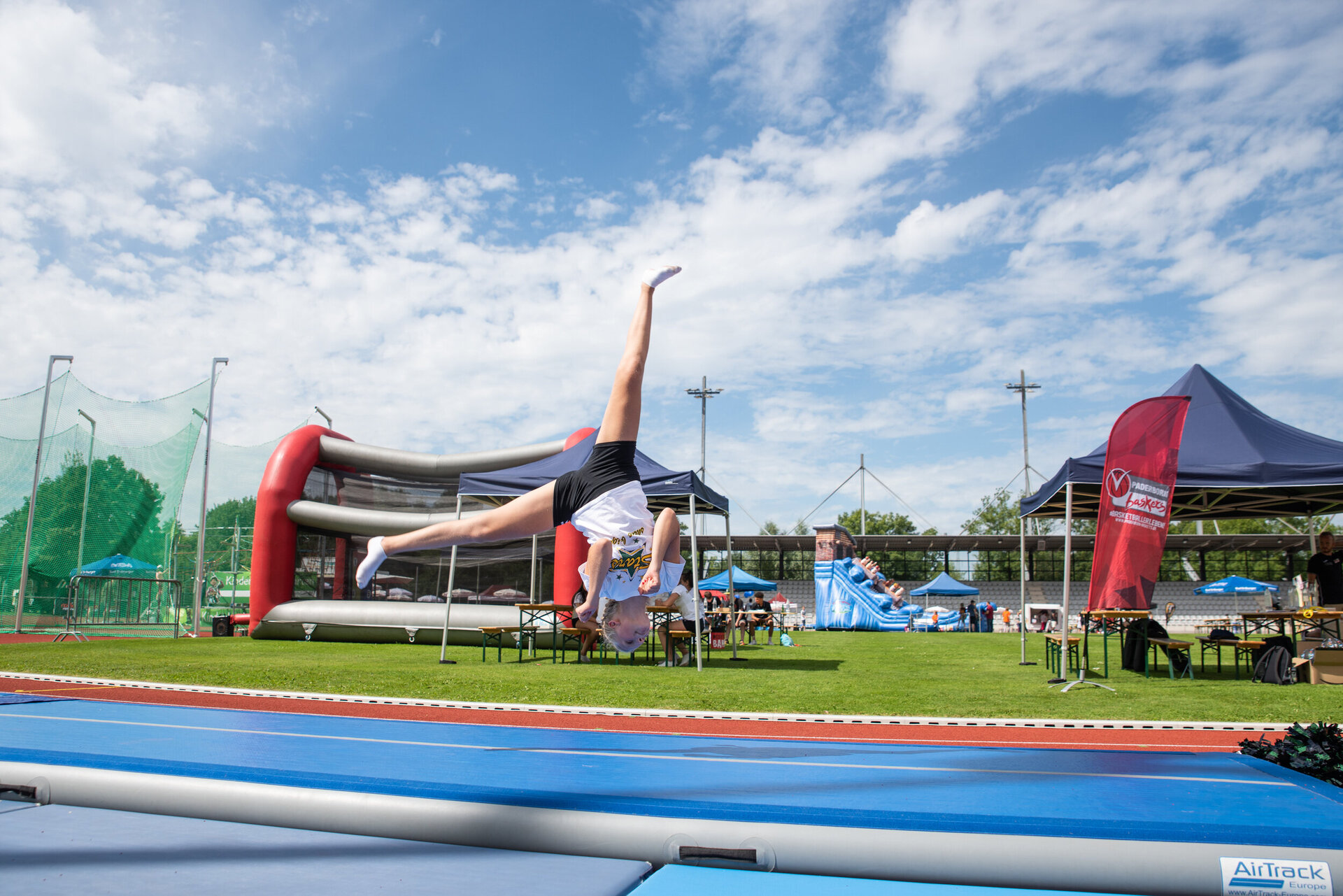Ein Kind macht einen Handstandüberschlag auf einer blauen AirTrack-Matte im Freien. Im Hintergrund sind auf einer Wiese Zelte, Sportgeräte und aufblasbare Spielstationen zu sehen, der Himmel ist teils bewölkt.