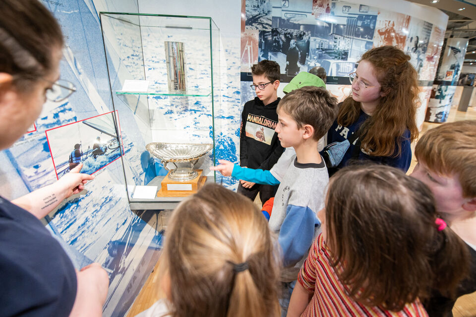Eine Gruppe von Schülern steht im Heinz Nixdorf MuseumsForum vor einer Vitrine mit einem großen silbernen Pokal und weiteren Ausstellungsstücken. Eine erwachsene Person zeigt auf ein Foto an der Wand, das Segler auf einem Boot zeigt, während die Kinder interessiert hinschauen.
