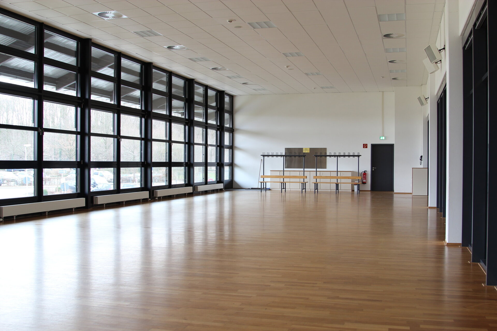 Großer, leerer Gymnastikraum mit hellem Holzfußboden, breiten Fensterfronten auf der linken Seite und Garderobenbänken mit Haken an der Rückwand.