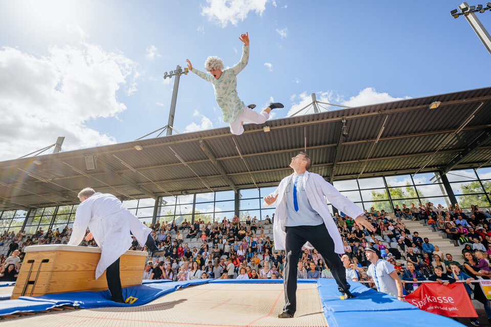 Eine große Zuschauertribüne ist gut gefüllt. Auf einer Trampolinfläche führen mehrere Personen in weißen Kitteln eine Show auf. Im Vordergrund springt eine als ältere Dame verkleidete Person hoch durch die Luft, während die anderen Akteure darunter stehen und das Publikum gebannt zuschaut.