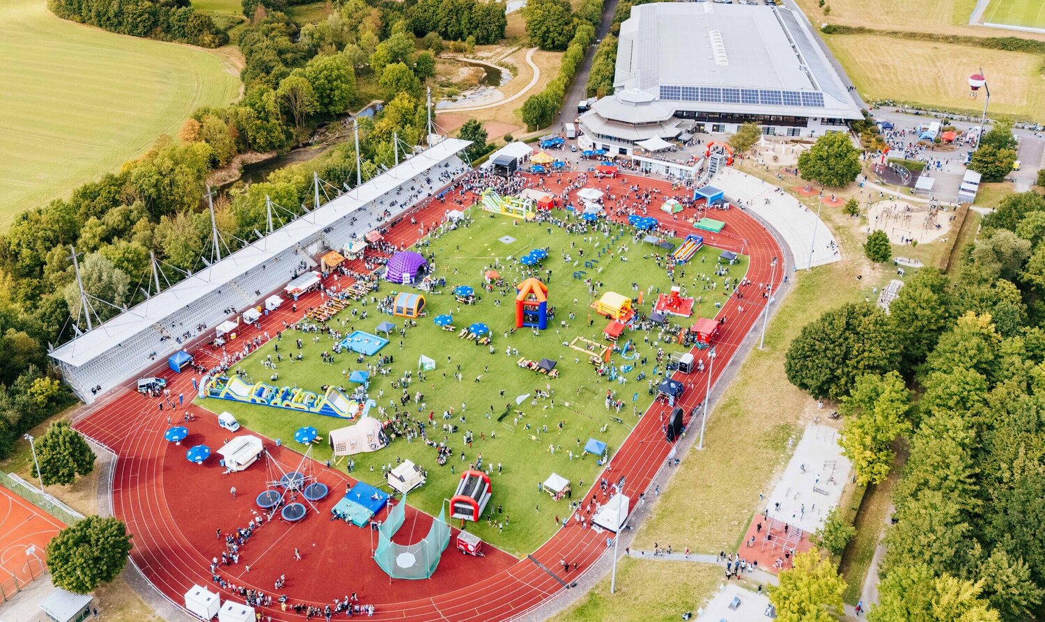 Luftaufnahme des Ahorn-Sportparks während des Radio Hochstift-Ferienfinales. Auf dem großen Sportfeld sind zahlreiche bunte Hüpfburgen, Spiel- und Mitmachstationen sowie Stände aufgebaut. Viele Menschen bewegen sich zwischen den Attraktionen, die Tribünen und umliegenden Wege sind ebenfalls belebt.