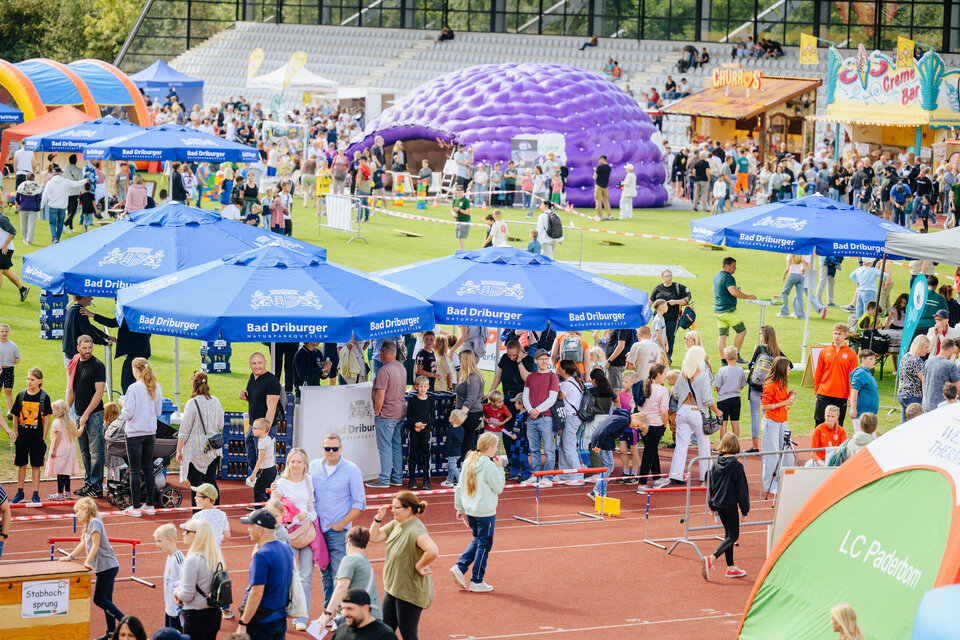 Menschen schlendern über das Festgelände beim Radio Hochstift-Ferienfinale im Ahorn-Sportpark. Unter blauen Sonnenschirmen stehen Besucher an Getränkeständen an. Dahinter bunte Stände, Hüpfburgen und ein großes lila Luftzelt.