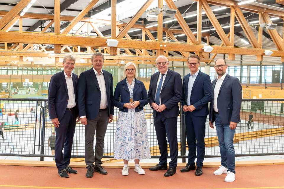 Bundestagsabgeordnete und Staatsministerin Dr. Christiane Schenderlein (Mitte) steht gemeinsam mit fünf politischen Vertretern im Ahorn-Sportpark Paderborn. Die Gruppe posiert in einer Reihe vor der großen Holz-Dachkonstruktion der Sporthalle. Von rechts: Prof. Dr. Nikolaus Risch, Martin Nixdorf, Bernhard Hoppe-Biermeyer, Dr. Carsten Linnemann und Ralf Pahlsmeier.