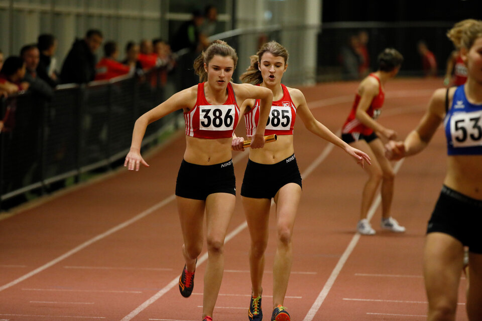 Bei einem Leichtathletik-Hallen-Wettkampf übergibt eine Läuferin den Staffelstab an die nächste Läuferin. Die Läuferinnen tragen rote Trikots mit den Startnummern 385 und 386. Im Hintergrund verfolgen zahlreiche Zuschauer den Wettkampf.