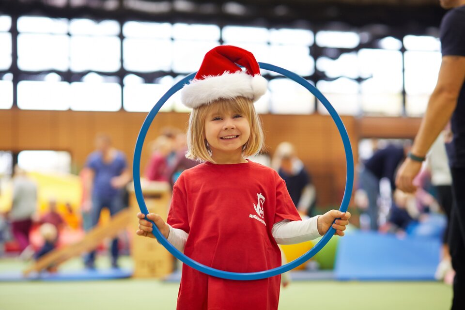 Ein Kind mit einem roten T-Shirt und einer Nikolausmütze steht im Vordergrund mit einem Reifen in der Hand. Durch den Reifen schaut das Kind in die Kamera. Im Hintergrund sind mehrere Personen an einem Sportgeräteaufbau angedeutet.