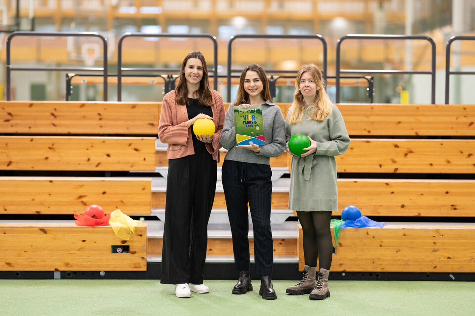 Das Organisationsteam für das bunte Sportprogramm für Familien stehen vor einer Tribüne in der Ahorn-Sportpark-Halle und halten Materialien in der Hand. Die linke Dame hat einen gelben Ball, die mittlere Dame das Kinderbuch "Vier-Farben-Land" und die rechte Dame einen grünen Ball in der Hand.