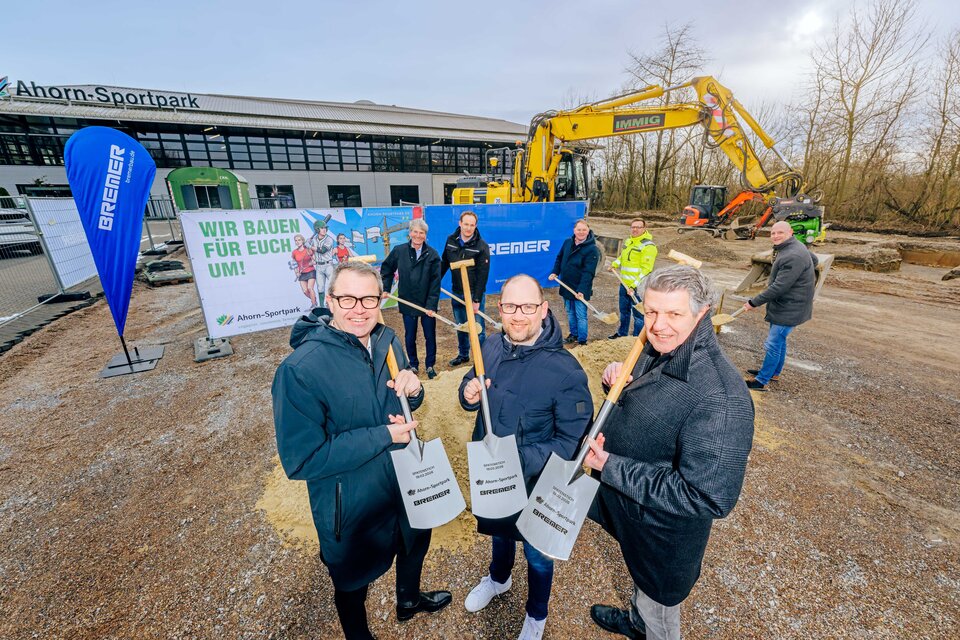 Das Foto zeigt den offiziellen Spatenstisch für den Start der Bauphase des neuen Kraft- und Athletikbereichs des Ahorn-Sportparks auf dem Nordparkparkplatz. Insgesamt acht Personen halten dabei einen Spaten in der Hand. Fünf Personen stechen mit ihrem Spaten in einen Haufen von Sand. Hinter der Personengruppe sind Bauzäune mit Banner augestellt sowie steht ein großer gelber Bagger. Ganz im Hintergrund ist das Ahorn-Sportpark-Gebäude zu erkennen.