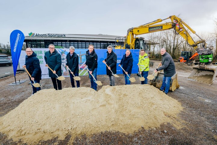 Das Foto zeigt den offiziellen Spatenstisch für den Start der Bauphase des neuen Kraft- und Athletikbereichs des Ahorn-Sportparks auf dem Nordparkparkplatz. Insgesamt acht Personen halten dabei einen Spaten in der Hand und stehen hinter einem Berg von Sand. Hinter der Personengruppe sind Bauzäune mit Banner augestellt sowie steht ein großer gelber Bagger. Ganz im Hintergrund ist das Ahorn-Sportpark-Gebäude zu erkennen.
